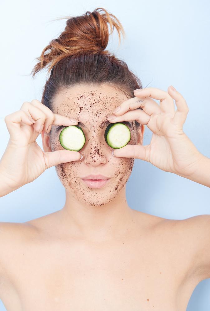 Woman with exfoliator on her skin holding cucumbers in front of her eyes
